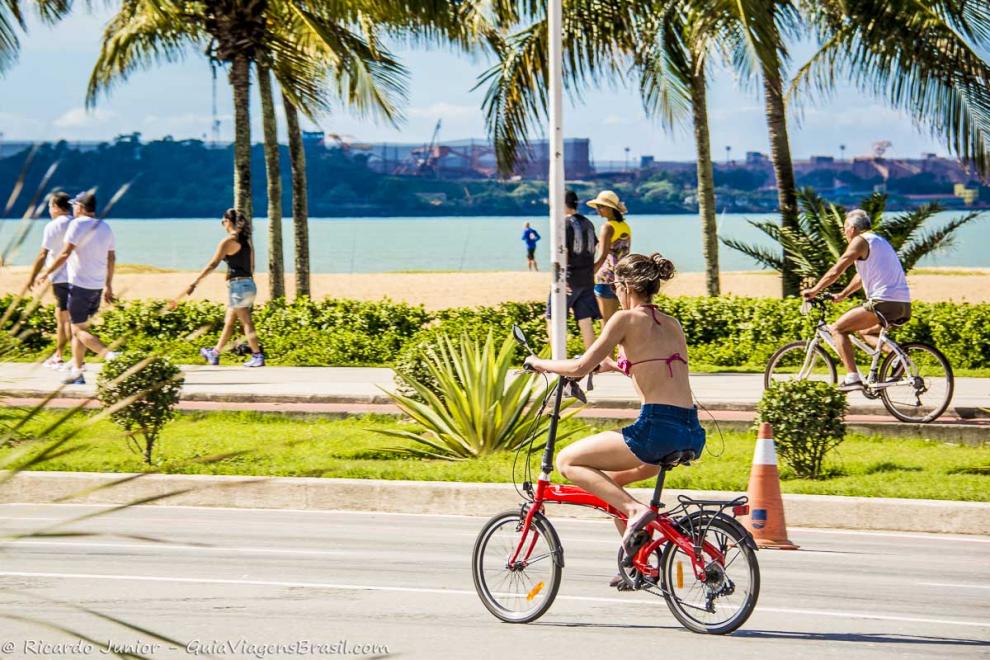 Imagem de uma moça de bicicleta na avenida e pessoas caminhando na ciclovia.